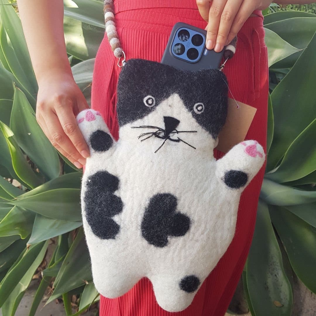 Black-and-white felted cat-shaped bag with spots, pink paw pads and a felt-ball strap
